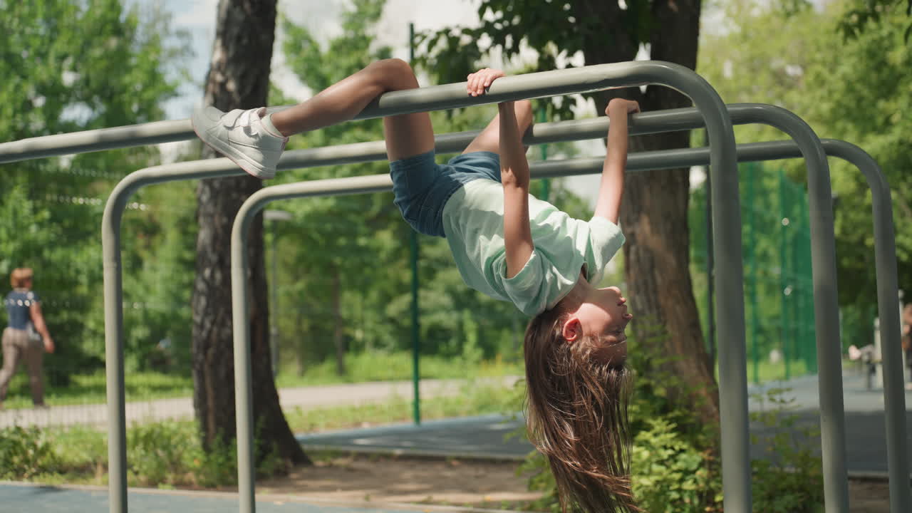 White daughter hanging across park bars, middistance shot of athletic girl performing inverted hang on metal rails, curly hair falling, sunny urban green space and playful strength