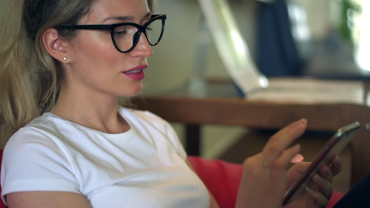 Blonde woman wearing black reading glasses and a white T-shirt, scrolling through her phone on a beanbag