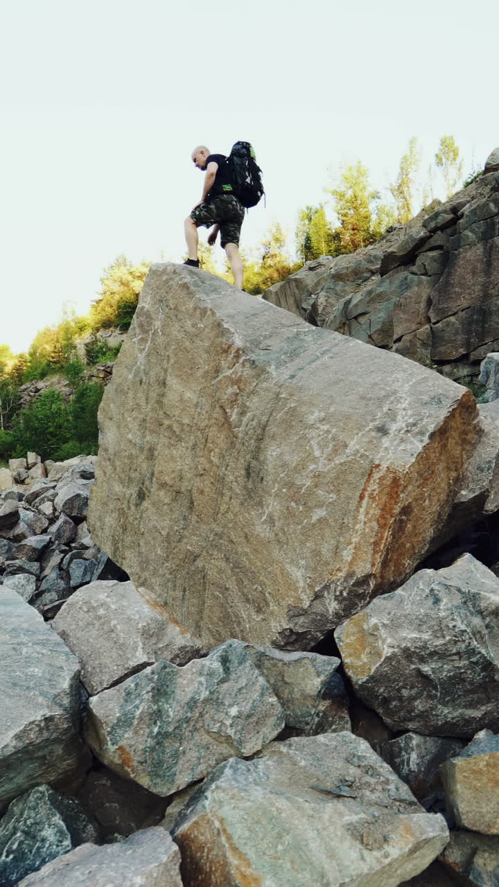 A traveler in shorts and with a backpack climbs onto a huge stone and sits on it amid a quarry with a lake in the center. Vertical video