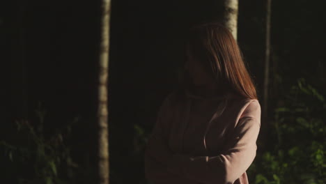 depressed young woman in warm pink coat peers thoughtfully into dark forest. lady thinks about problems and remembers past while enjoying nature near lighter