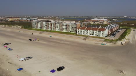 Elevándose-Sobre-La-Playa-En-Diagonal-Apuntando-Hacia-El-Alojamiento,-Mostrando-Vehículos-Estacionados-En-La-Playa,-Varios-Ingresando-A-La-Carretera-De-Acceso-A-La-Playa