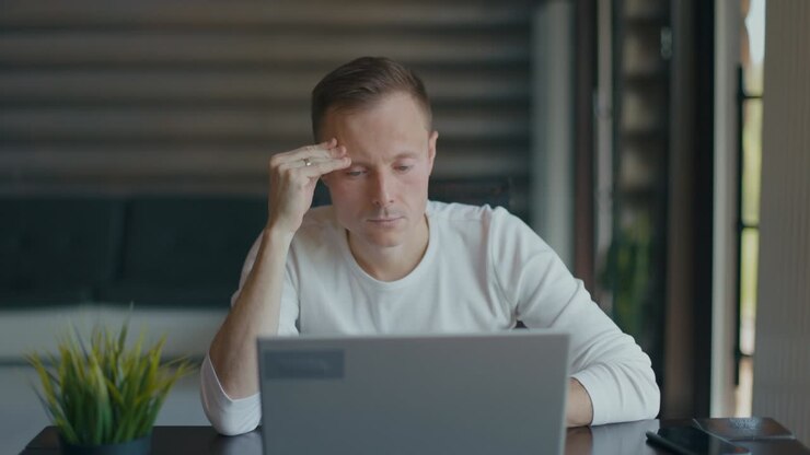Un giovane concentrato è seduto alla scrivania nella stanza con il laptop. Si strofina la fronte con la mano destra.