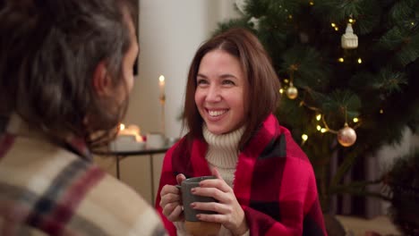 Over-the-shoulder,-a-brunette-guy-in-a-checkered-shirt-is-talking-to-his-brunette-girlfriend-in-a-white-sweater-and-a-red-checkered-shirt-who-is-drinking-a-hot-drink-from-a-gray-mug-near-a-green-New-Year-tree-in-a-cozy-room-in-winter