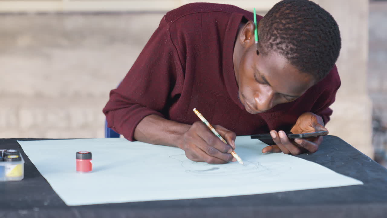 Black man painting on paper, dynamic action shot of young African artist applying ink and brush, small paint jar and smartphone reference on table, outdoor workshop vibe, intense concentration