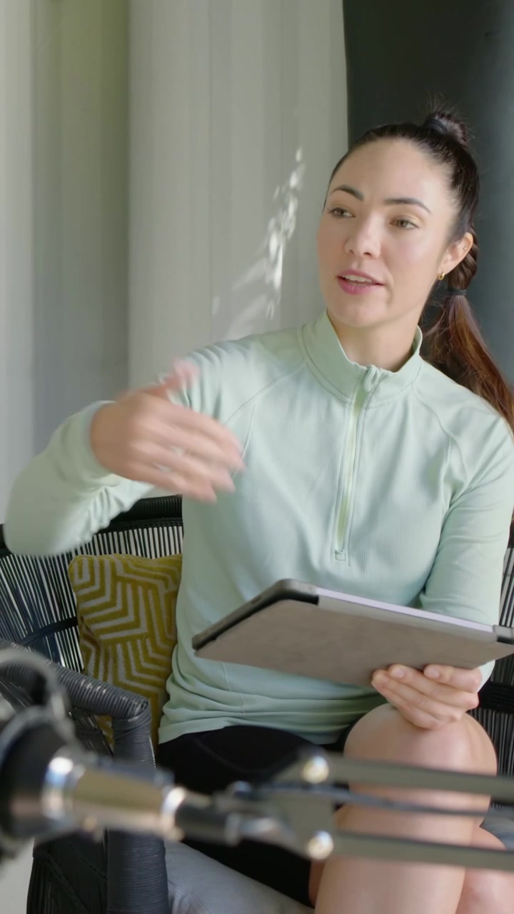 Video verticale: Donna con top color menta e pantaloncini neri, che legge un tablet e conduce una chat in poltrona, con un microfono a giraffa.