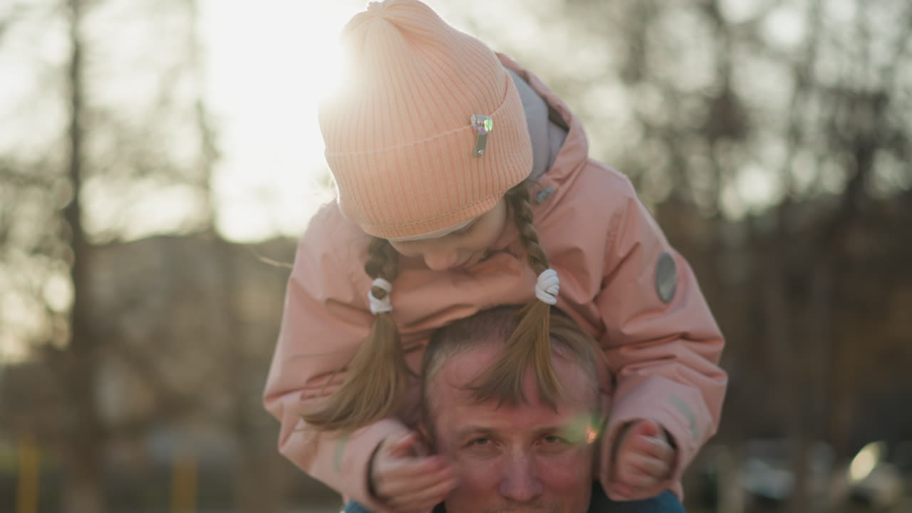 Sweet moment in the park as a little girl in a pink cap covers her father's eyes while sitting on his shoulders, both enjoying the playful interaction