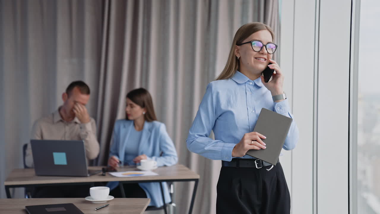 Business lady in glasses speaks on the phone standing in the office at the window. Woman is holding paper notebook in her hand. Colleagues working at backdrop.