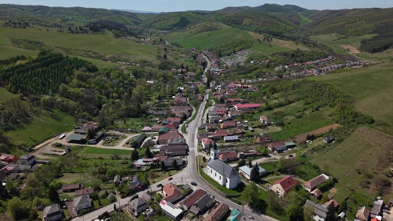 Una vista aerea cattura un pittoresco villaggio europeo immerso in una lussureggiante valle verde, circondato da colline verdeggianti e una strada principale tortuosa.