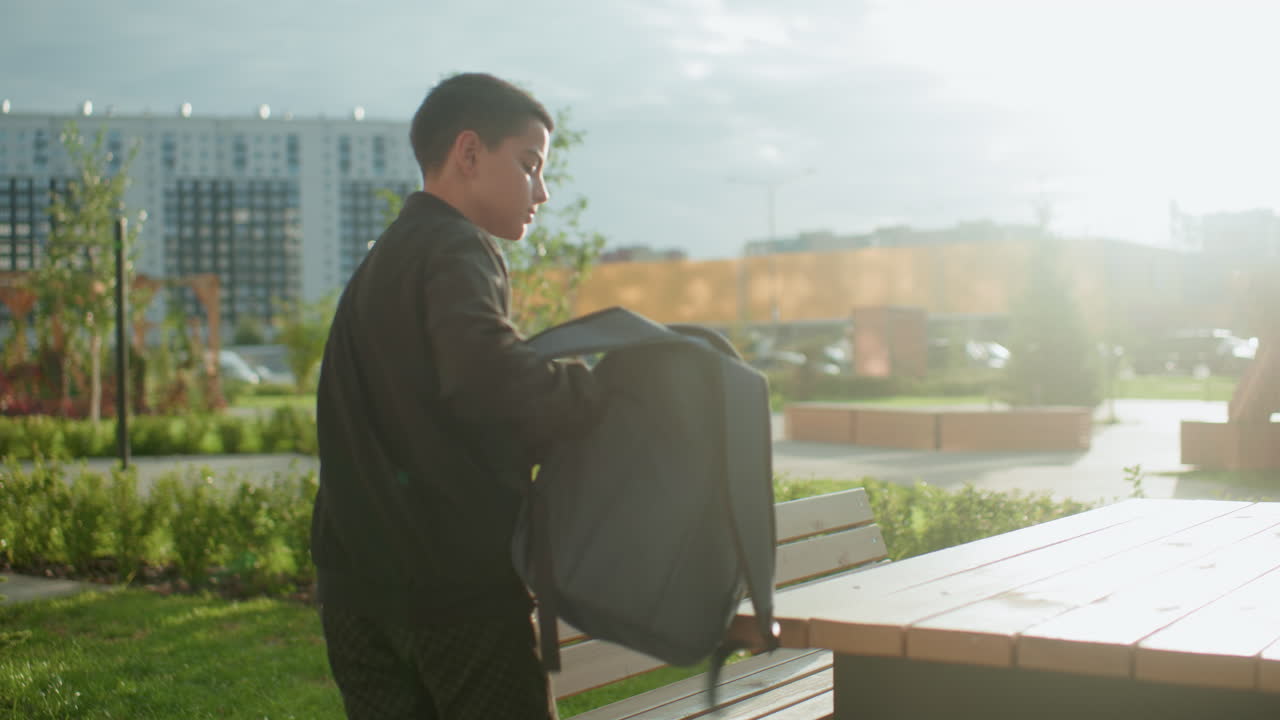 Young boy carrying backpack walks toward bench under warm daylight, preparing to drop his bag on wooden seat, blur background of city buildings and greenery visible with natural light glow