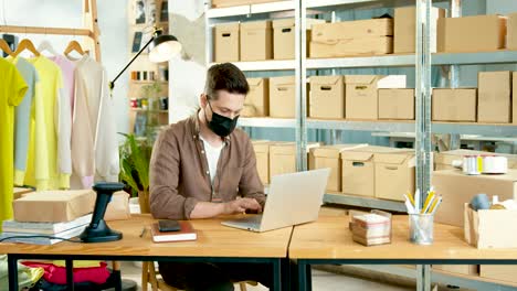 Caucasian-designer-man-wearing-black-mask-working-on-laptop-while-sitting-at-desk-in-clothing-shop-warehouse