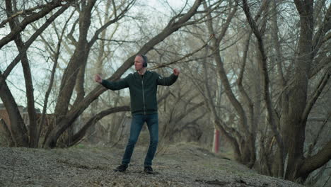 a man in a green jacket with headphones dances alone in a serene forest, the ground is covered with leaves and twigs