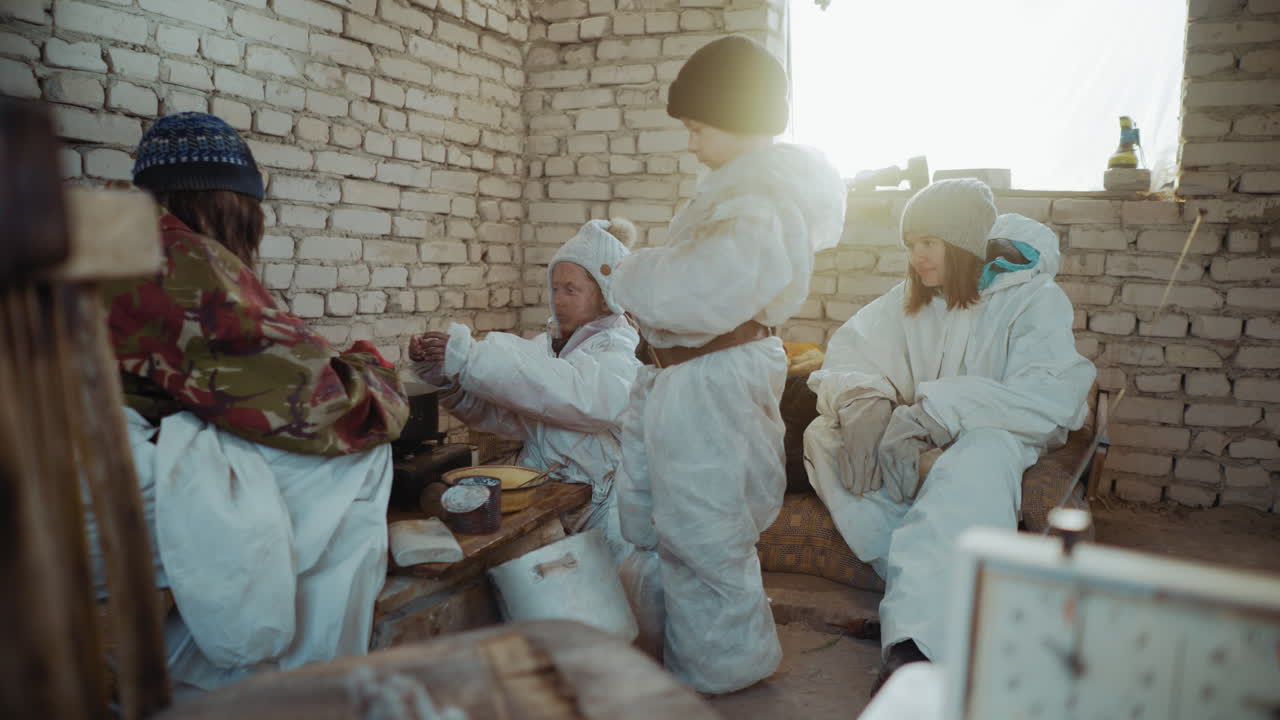 Survivors in protective clothes gather inside ruined brick shelter, child stands near stove as others sit waiting, cold sunlight streams through window showing hardship of survival after disaster