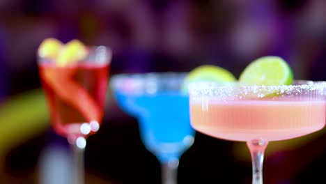 three colorful cocktails with garnishes in a lively bar environment, featuring bright lighting and a festive atmosphere