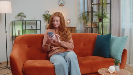 happy young woman sitting on sofa using smartphone texting smiling at home living room apartment