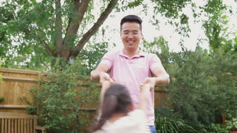 asian father swinging daughter by arms as they play game in garden together