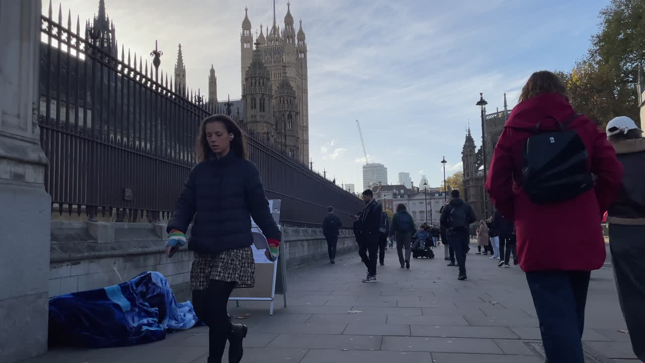 People walk past a sleeping homeless person underneath a blue blanket in front of the Houses of Parliament and the Victoria Tower on a clear winter morning.
