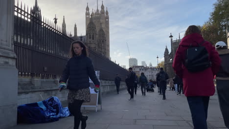 people walk past a sleeping homeless person underneath a blue blanket in front of the houses of parliament and the victoria tower on a clear winter morning.