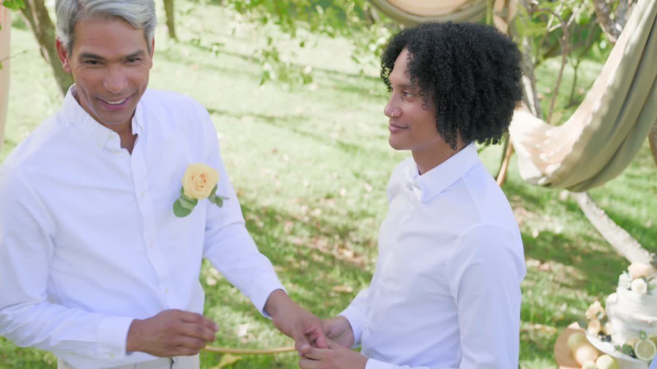 Partner uomini che si scambiano le fedi nuziali durante una cerimonia di matrimonio in un giardino sotto un arco di tessuto drappeggiato.