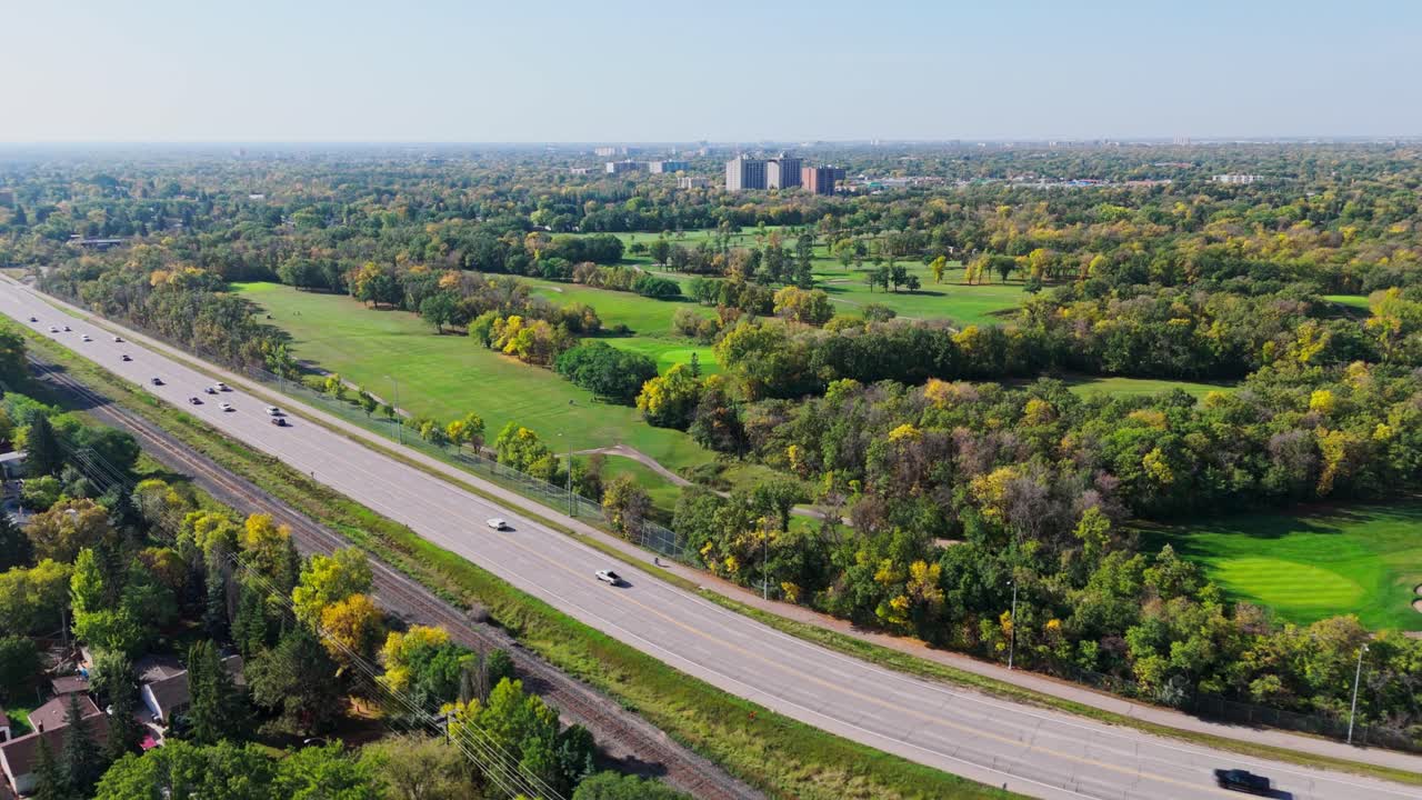 Drone footage of vehicles on a highway next to a golf course surrounded by green parkland and autumn trees. City buildings visible in the distance