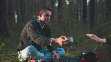 man sits cross legged on grass in forest reaching for canned food near boiling pot as sunlight filters through trees during serene outdoor camping experience