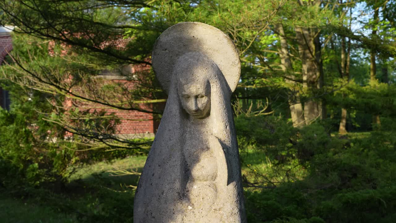 A close-up shot of a stone statue representing the Virgin Mary, located in the Central Cemetery in Szczecin. Surrounded by greenery and bathed in soft light. Shot handheld, no stabilization used.