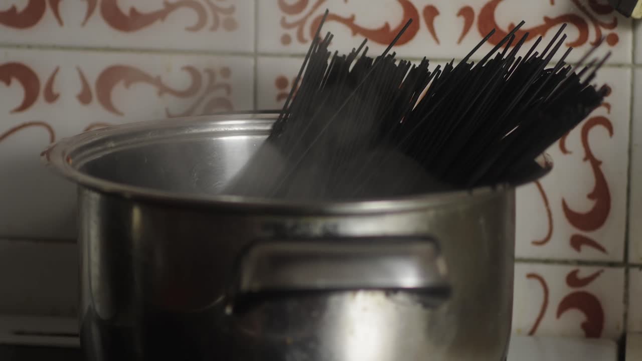 Pot filled with uncooked black spaghetti placed on cooker against a tiled wall with ornate designs