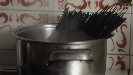 pot filled with uncooked black spaghetti placed on cooker against a tiled wall with ornate designs
