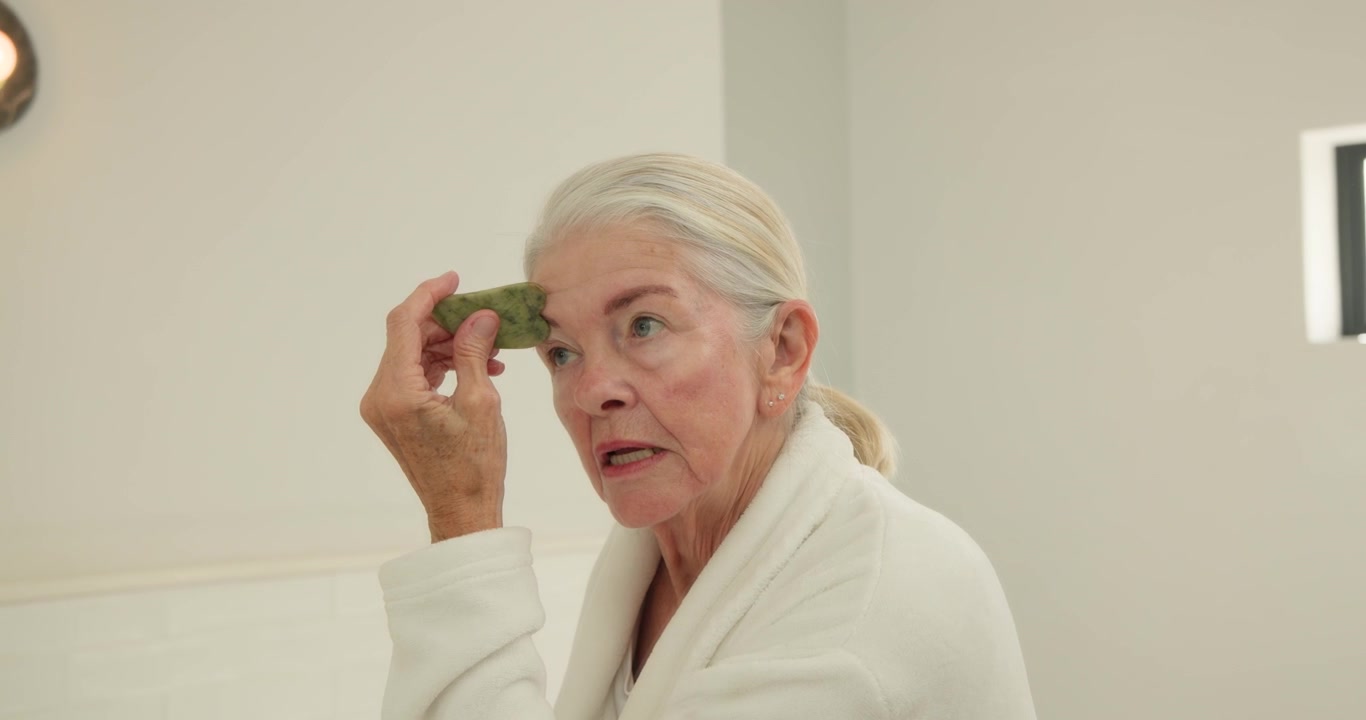 Donna anziana nel bagno di casa che indossa un accappatoio, posiziona una pietra verde sull'occhio destro e si massaggia la fronte.