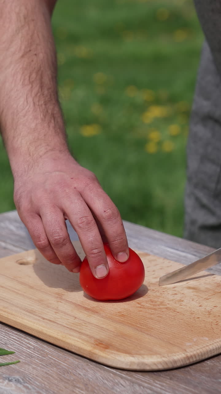 Man cutting tomato on a board outdoors. Preparing dinner at dacha. Man cuts vegetable into four parts on a table before the picnic. Vertical video