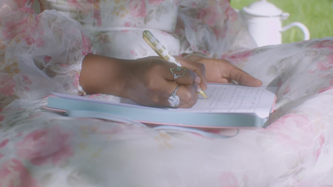 Black lady with floral dress sits down and writes in diary, close up dolly