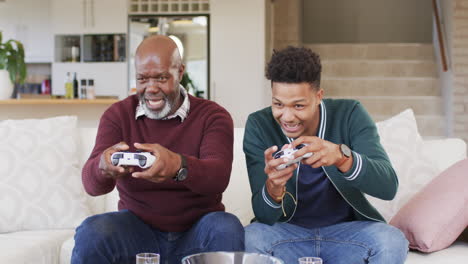 excited african american father and adult son playing video game with gamepads at home, slow motion