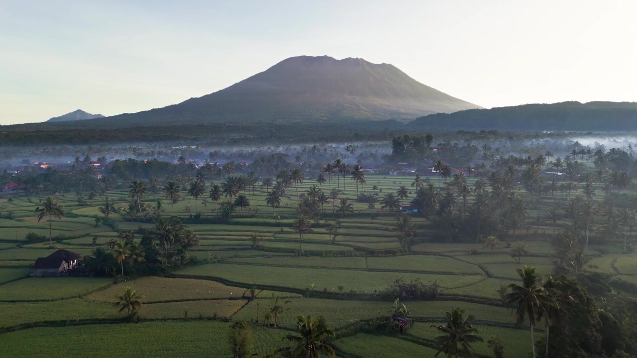 Vulcano Monte Agung che si erge sopra risaie e palme con un villaggio rurale nella morbida luce del mattino e nella foschia attraverso l'ampio paesaggio agricolo di Bali, Indonesia, filmato con drone in volo in avanti.