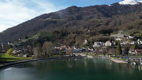 Drone-clip-showing-view-of-traditional-white-buildings-on-shore-of-calm-lake,-with-snow-topped-mountains-in-the-background