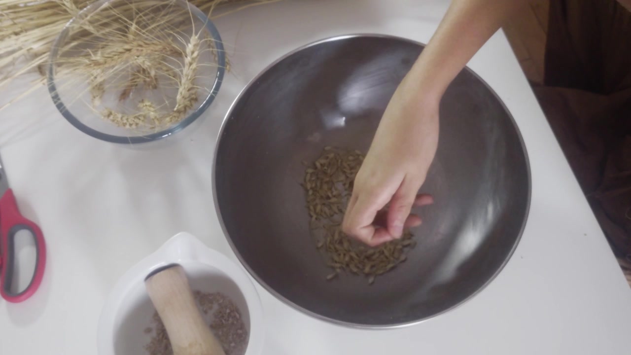 A high-angle overhead shot shows a hand carefully sorting through harvested golden wheat grains inside a stainless steel bowl to remove remaining husks on a white table during a grain processing tas