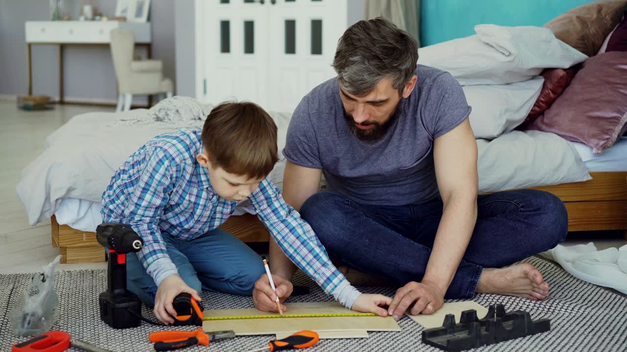 padre e hijo están midiendo una pieza de madera con un carrete de medición preparándose para construir algo juntos dentro de la casa. diferentes herramientas y muebles modernos son visibles.