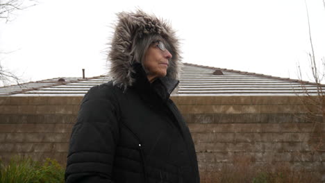 una anciana con una gruesa chaqueta de invierno y una capucha parada en una helada tormenta de nieve de invierno a cámara lenta