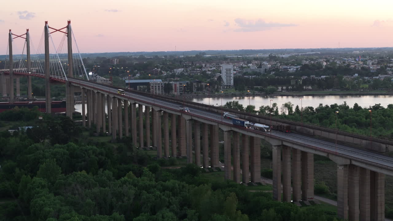 Aerial shot of a large cable-stayed bridge with trucks and cars crossing over a river, leading to a city skyline at sunset. A scene of modern transportation.