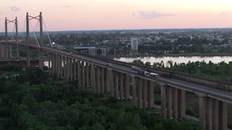 aerial shot of a large cable-stayed bridge with trucks and cars crossing over a river, leading to a city skyline at sunset. a scene of modern transportation.