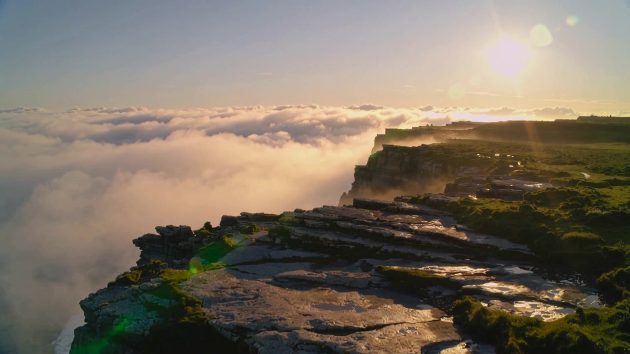 Un dron que se acerca revela el borde de un acantilado costero, losas y charcos que reflejan el sol y un mar de nubes.
