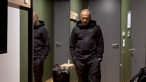 bald young indian male pacing past large mirror in hallway talking wirelessly dressed in black trousers and hoody