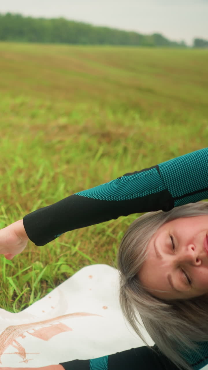 요가 매트에 한 쪽을 누워있는 은 여자가 팔을 펴고 은 자세를 연습하고, 먼 곳의 자연과 나무로 둘러싸인 구름이 은 하늘 아래의 광활한 풀에서