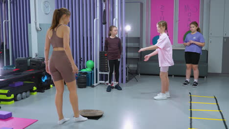 back view of an instructor observing a girl in pink demonstrating a learned exercise while two other kids watch, the instructor moves an object forward in a well-equipped gym