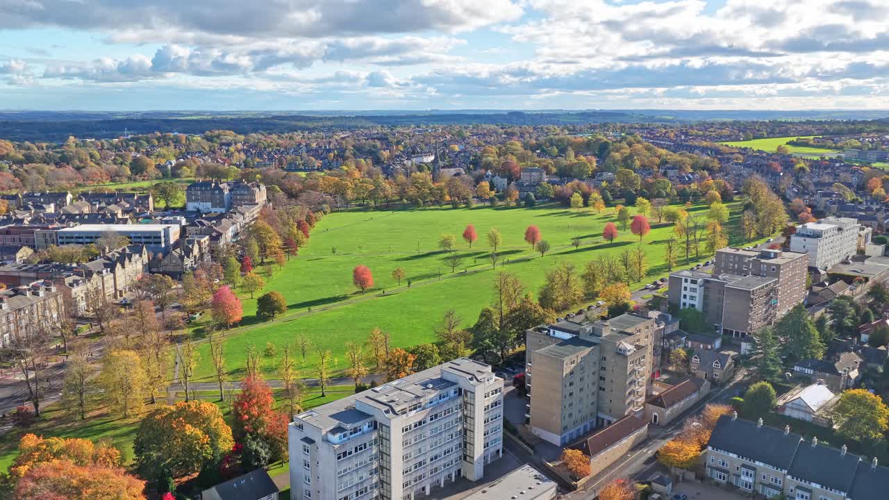 Drone view of Harrogate, North Yorkshire, England, showing autumn trees surrounding the large open parkland known as The Stray, with residential and historic town buildings under a blue clody sky