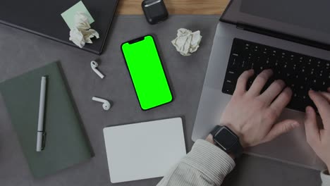 person typing on laptop with green screen phone and office supplies