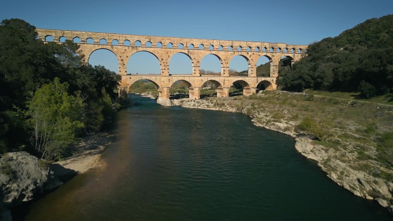 Ripresa con drone del Pont du Gard, un sito Patrimonio dell'Umanità in Francia. Un acquedotto romano che attraversa un fiume è illuminato dal sole serale. La struttura si riflette nell'acqua