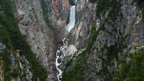 Ascending-top-down-shot-of-waterfall-and-river-between-gigantic-mountain-wall-in-Slovenia