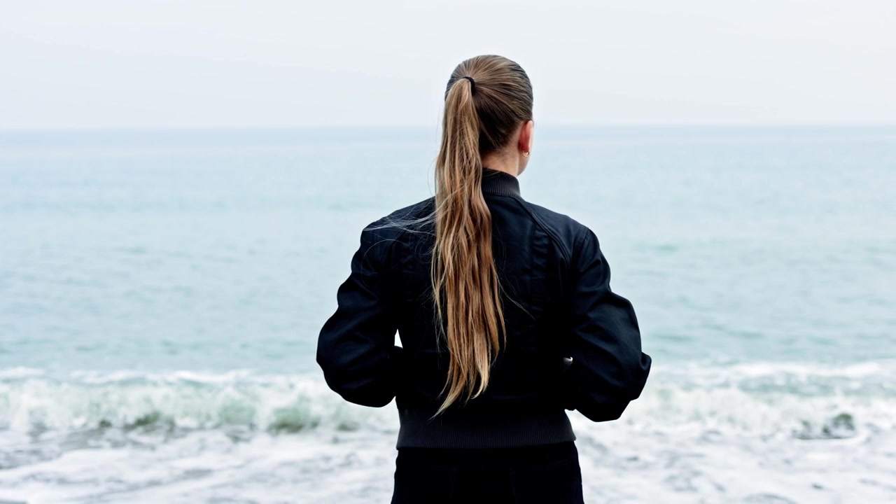 Young woman standing and looking at the sea, view from the back. Woman walking along the seashore. Girl with long hair walking near the ocean in autumn.