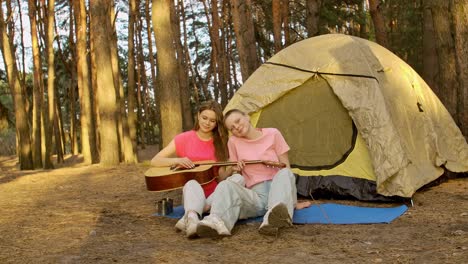 two friends playing guitar in a forest campsite