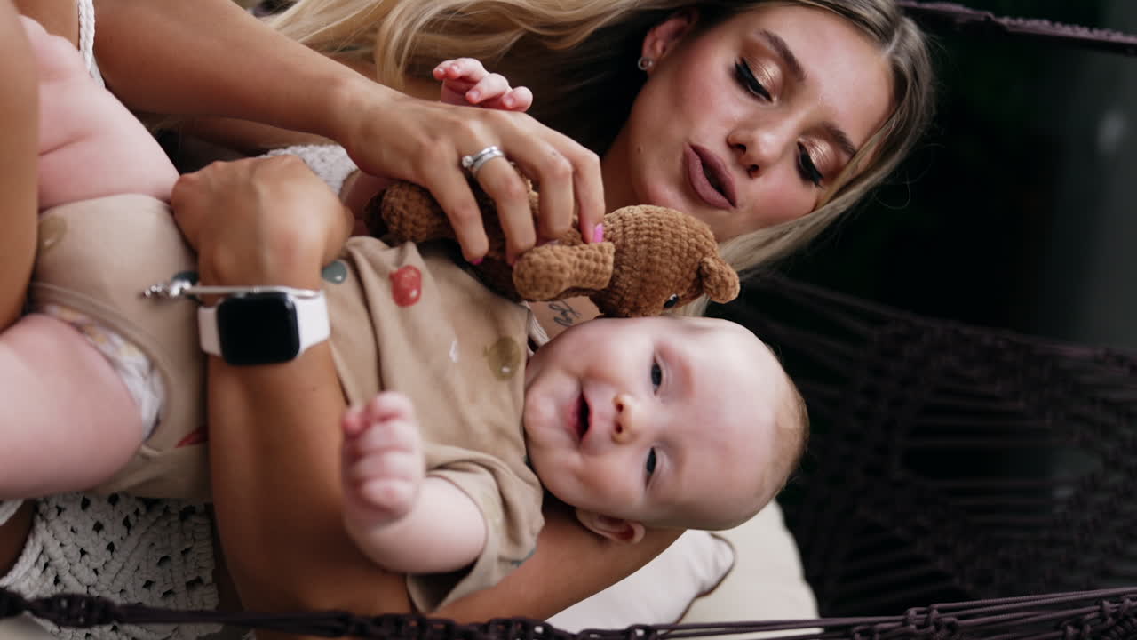 Beautiful blonde long-haired lady holding her baby in hands. Mother gives a brown knitted teddy to her child. Vertical screen.