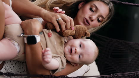 beautiful blonde long-haired lady holding her baby in hands. mother gives a brown knitted teddy to her child. vertical screen.